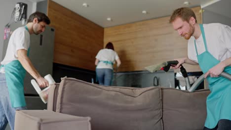 Confident-group-of-three-cleaners-in-a-white-T-shirt-and-blue-aprons-are-cleaning-a-modern-kitchen-in-an-apartment-on-call.-The-cleaning-company-sends-a-group-of-cleaners-to-clean-the-apartment