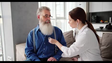 Una-Doctora-Morena-Con-Bata-Blanca-Examina-A-Un-Hombre-De-Cabello-Gris-Y-Barba-Exuberante-Con-Camisa-Azul-Usando-Un-Estetoscopio-Y-Aplica-El-Instrumento-Al-Pecho-Del-Hombre-En-Un-Apartamento-Moderno.