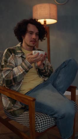 Vertical-video-of-a-brunette-guy-with-curly-hair-brings-a-cup-of-tea-to-his-face-with-both-hands-and-enjoys-a-hot-drink-at-home-on-a-chair-near-the-lamp