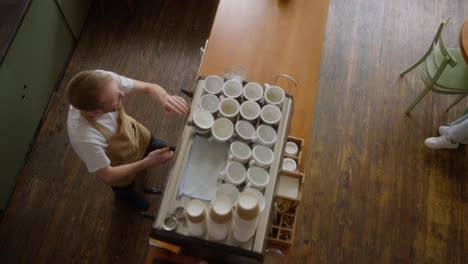 Top-view-of-happy-guy-blond-barista-in-light-brown-behind-coffee-machine-with-many-coffee-cups-near-counter-in-cafe