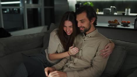 Happy-brunette-girl-has-fun-and-rejoices-hugging-her-brunette-husband-with-stubble-in-a-beige-shirt-while-he-works-remotely-using-a-gray-laptop-on-the-sofa-in-a-modern-apartment-in-the-evening