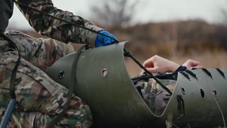 From-the-side-close-up-a-confident-male-army-medic-in-camouflage-green-clothing-and-blue-rubber-gloves-ties-straps-on-a-stretcher-to-move-a-wounded-soldier-during-combat-operations-in-the-steppe-outside-the-city