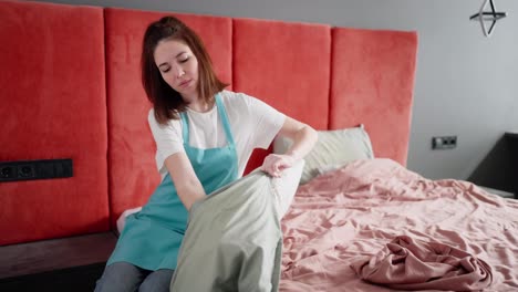 A-brunette-cleaning-lady-girl-in-a-white-T-shirt-and-a-blue-apron-tucks-a-white-pillow-into-a-gray-pillowcase-while-cleaning-and-making-the-bed-in-the-bedroom-of-a-modern-apartment