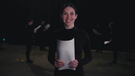 Portrait-of-a-happy-and-confident-young-brunette-girl-in-a-black-actress-costume-who-holds-sheets-of-script-and-character-text-in-her-hands-during-her-preparation-for-a-performance-in-the-theater