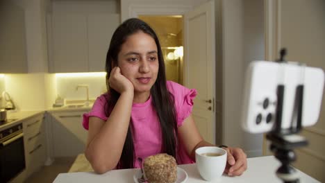 A-happy-brunette-girl-in-a-pink-dress-sits-at-a-table-in-front-of-her-with-a-small-cake-and-a-mug-of-white-coffee.-The-girl-communicates-via-video-with-her-viewers-and-friends-in-a-modern-apartment-at-home