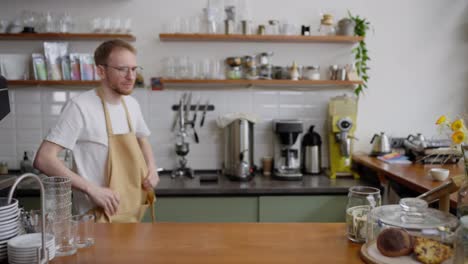 Portrait-of-a-confident-blond-guy-in-glasses-puts-on-a-yellow-apron-and-prepares-for-a-working-day-at-the-counter-in-a-cafe