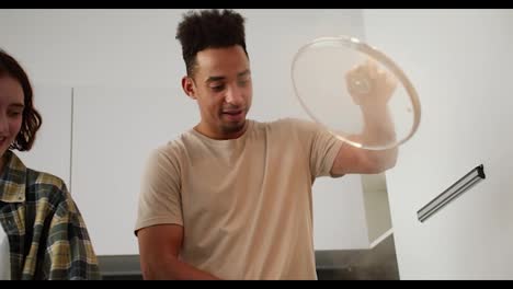 A-happy-young-man-with-Black-skin-in-a-beige-T-shirt-lifts-the-lid-of-a-frying-pan-and-a-large-amount-of-steam-comes-out-from-there-while-preparing-breakfast-with-his-young-adult-brunette-girlfriend-with-a-bob-hairstyle-in-a-modern-kitchen