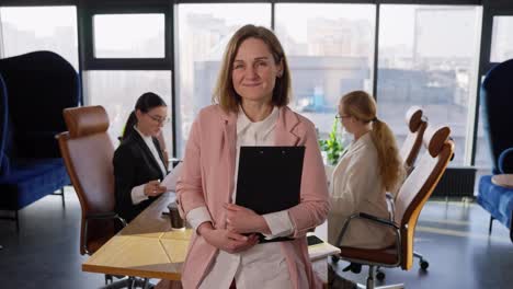 Portrait-of-a-happy-middle-aged-blonde-girl-in-a-pink-business-suit-with-a-tablet-in-her-hands-posing-near-the-table-in-a-modern-office-with-large-windows