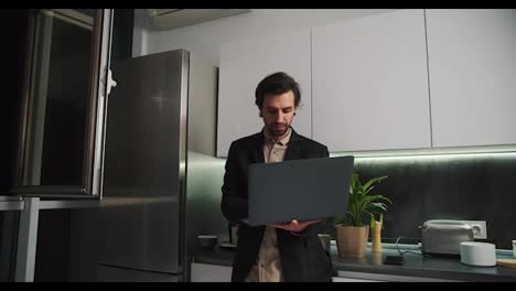 A-serious-brunette-man-with-stubble-in-a-black-jacket-and-beige-shirt-stands-holding-in-his-hands-a-gray-laptop-working-on-it-only-in-beige-shorts-in-a-modern-kitchen-in-an-apartment-while-working-remotely-in-the-evening