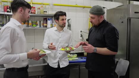 Dos-Camareros-Con-Camisas-Blancas-Se-Comunican-Con-Un-Chef-Con-Uniforme-Negro-Sobre-La-Calidad-De-La-Comida-Mientras-Están-En-La-Cocina-De-Un-Restaurante.-Un-Chef-Profesional-Con-Uniforme-Negro-Les-Dice-A-Los-Camareros-Cómo-Servir-Un-Plato-Correctamente.