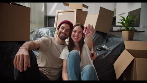 Portrait-of-a-happy-brunette-girl-in-a-white-T-shirt-who-holds-the-keys-in-her-hand-and-shows-them-while-sitting-on-the-floor-with-her-brunette-boyfriend.-In-a-beige-T-shirt-near-the-sofa-there-is-a-packed-plastic-case-and-a-huge-number-of-boxes-around-in-a-new-apartment