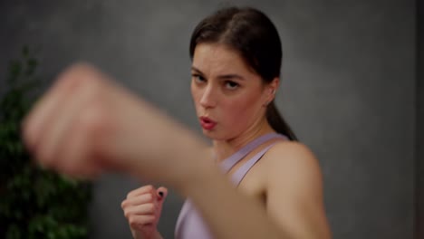 Close-up-dynamic-video-portrait-of-a-confident-brunette-girl-in-a-purple-top-who-practices-boxing-strikes-in-a-modern-apartment-while-playing-sports-at-home