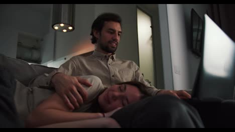 Close-up-of-a-happy-brunette-girl-in-a-beige-T-shirt-sleeping-on-the-lap-of-her-boyfriend-who-is-working-on-a-Gray-laptop-in-the-evening-remotely-at-home-in-a-modern-apartment