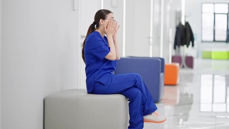 Sad-girl-brunette-doctor-in-round-glasses-and-a-blue-uniform-sits-on-a-soft-chair-in-a-modern-corridor-in-the-clinic-and-is-sad-about-failure-while-working