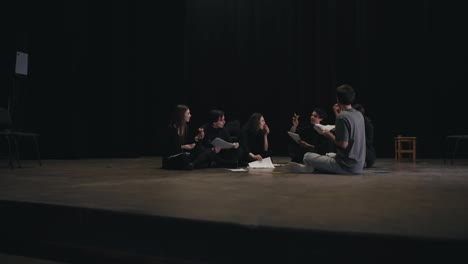 A-group-of-young-confident-actors-in-black-uniforms-are-talking-with-the-director-about-the-script-for-their-next-play-and-are-sitting-on-stage-in-the-theater