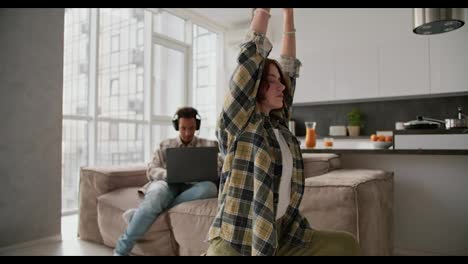 A-girl-with-brown-hair-and-a-bob-hairstyle-in-a-green-checkered-shirt-does-yoga-and-sports-while-her-boyfriend-a-young-man-with-Black-skin-in-black-headphones-works-at-his-Gray-laptop-Sitting-on-the-sofa-in-a-modern-studio-apartment