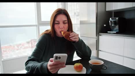 Happy-and-confident-brunette-girl-in-a-green-sweater-eats-a-croissant-and-tea-during-her-breakfast-and-surfs-social-networks-in-her-white-smartphone-in-a-modern-apartment-in-the-kitchen