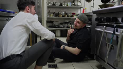 Un-Cocinero-Cansado,-Con-Uniforme-Negro-Y-Barba,-Le-Cuenta-A-Un-Joven-Camarero-Con-Camisa-Blanca-Lo-Duro-Que-Fue-Su-Jornada-Laboral-Y-El-Camarero-Apoya-Al-Hombre-Mientras-Trabaja-En-La-Cocina-De-Un-Restaurante.