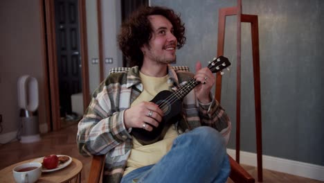 Happy-brunette-guy-in-a-checkered-shirt-with-curly-hair-plays-the-ukulele-and-sits-on-a-chair-in-a-cozy-modern-apartment