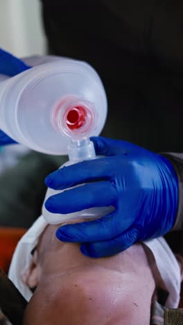 Vertical-video-close-up-of-a-confident-man-in-blue-medical-gloves-pumping-air-and-performing-artificial-respiration-on-a-man-who-is-lying-on-a-couch-in-a-small-medical-room