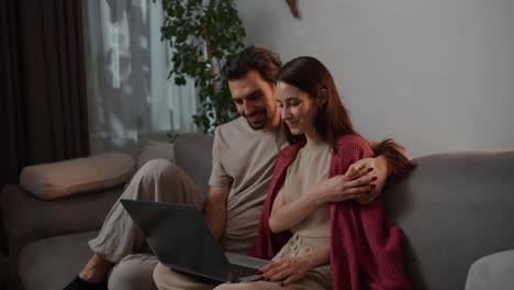 Side-view-of-a-happy-brunette-man-with-stubble-together-with-his-young-brunette-wife-in-a-red-sweater-sitting-on-a-gray-sofa-and-watching-an-interesting-movie-on-a-gray-laptop-in-a-modern-apartment