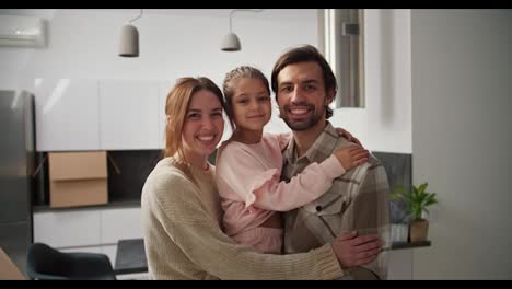 Portrait-of-a-happy-family-after-moving-a-brunette-man-with-stubble-holds-his-little-daughter-in-pink-clothes-in-his-arms-and-they-are-hugged-by-a-brunette-girl-in-a-beige-sweater-while-in-a-modern-apartment-after-moving