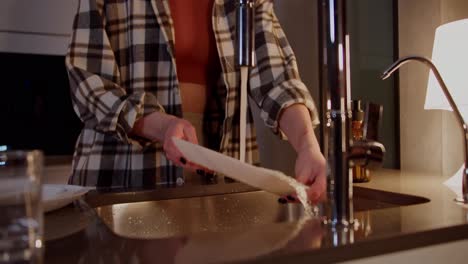 Close-up-shot-of-a-happy-and-cheerful-girl-in-a-checkered-shirt-listening-to-music-on-wireless-red-headphones-and-washing-dishes-in-the-kitchen-in-a-modern-apartment-in-the-evening