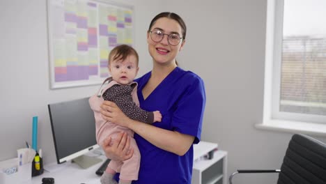 Portrait-of-a-confident-and-happy-brunette-doctor-girl-in-glasses-and-a-blue-uniform-who-is-holding-a-little-baby-girl-in-her-arms.-Portrait-of-a-confident-and-qualified-female-doctor-in-a-blue-pediatrician-uniform-in-a-modern-clinic