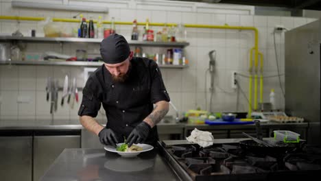 Confident-professional-male-chef-in-a-black-uniform-puts-dishes-on-a-plate-and-decorates-it-with-greens-before-serving-to-the-hall.-Male-chef-serving-dishes-and-adding-finishing-touches-before-serving-in-a-restaurant