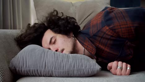 A-tired-brunette-guy-with-curly-hair-in-a-checkered-shirt-falls-on-a-gray-pillow-at-home-after-a-hard-day-at-work