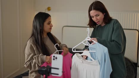 A-happy-brunette-girl-in-a-gray-sweater-together-with-her-friend-in-a-green-sweater,-talk-about-comfortable-summer-colorful-clothes-while-maintaining-her-Beauty-blog-in-a-modern-apartment