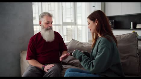 An-elderly-man-with-gray-hair-and-a-lush-beard-in-a-red-T-shirt-measures-his-blood-pressure-together-with-his-daughter-a-brunette-adult-girl-in-blue-pants-and-a-green-jacket-sitting-on-a-brown-sofa-in-a-modern-apartment