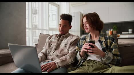 Happy-young-Black-skinned-brunette-man-with-stubble-in-a-checkered-cream-shirt-sits-with-his-girlfriend-and-looks-at-something-on-his-gray-laptop-while-spending-time-together-on-the-sofa-in-a-modern-studio-apartment