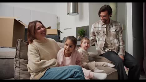 Happy-family-on-the-sofa-after-moving-to-a-new-modern-apartment-communicates.-A-brunette-man-with-stubble-in-a-checkered-beige-shirt-his-brunette-wife-their-little-daughter-in-pink-clothes-and-their-son-are-sitting-on-the-sofa-after-moving-into-a-new-modern-apartment