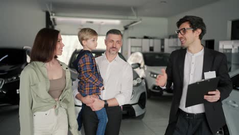 Close-up-a-confident-male-brunette-assistant-with-glasses-in-a-business-suit-with-a-tablet-in-his-hands-tells-the-family-about-what-cars-are-in-the-car-dealership-and-about-modern-car-models