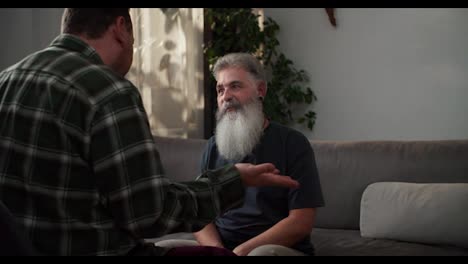 Un-Hombre-Feliz-Con-Cabello-Gris-Y-Una-Exuberante-Barba-Gris-Se-Divierte-Y-Se-Regocija-Durante-Su-Comunicación-Con-Su-Novio,-Una-Morena-Con-Barba-Incipiente-Con-Una-Camisa-A-Cuadros-Sentada-En-Un-Sofá-Gris-En-Un-Apartamento-Moderno.