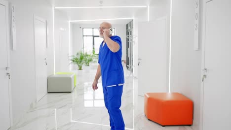 Confident-male-doctor-of-advanced-age-in-glasses-with-a-gray-beard-in-a-blue-uniform-communicates-on-a-white-smartphone-and-looks-around-while-standing-in-the-corridor-of-a-modern-clinic