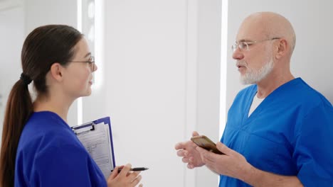 Ein-Selbstbewusster-Arzt-Mit-Brille-Und-Weißem-Bart-In-Blauer-Uniform-Kommuniziert-Während-Seiner-Pause-Auf-Dem-Flur-Einer-Hellen,-Modernen-Klinik-Mit-Seiner-Kollegin,-Einem-Brünetten-Mädchen-Mit-Runder-Brille