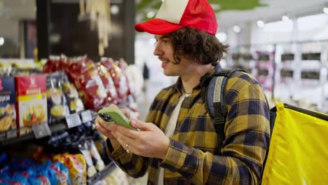 A-confident-food-delivery-guy-in-a-checkered-shirt-walks-near-the-aisles-in-a-supermarket-and-chooses-the-products-he-needs-reading-them-on-his-phone