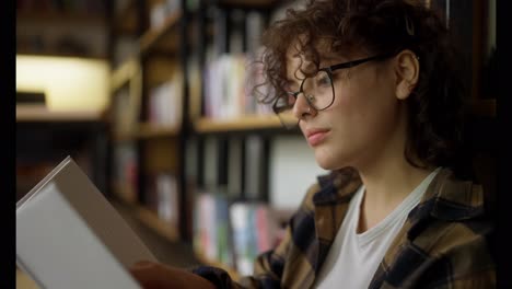 Close-up-an-interested-girl-with-curly-hair-and-glasses-sits-near-shelves-with-books-and-reads-one-in-the-library