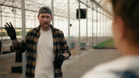 Over-his-shoulder-a-confident-guy-in-a-cap-with-a-beard-in-a-checkered-shirt-is-talking-to-a-girl-farmer-and-discussing-plans-for-work-in-a-greenhouse-on-the-farm