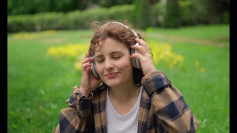 Close-up-of-a-happy-and-cheerful-girl-with-curly-hair-in-a-white-T-shirt-puts-on-wireless-headphones-and-listens-to-music-on-the-green-grass-in-the-park