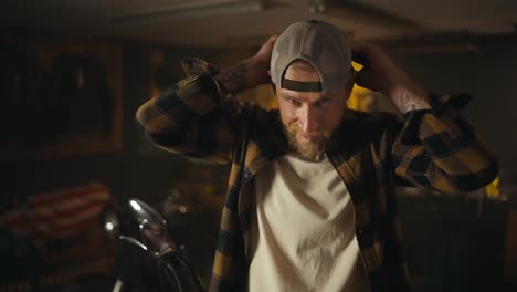 Portrait-of-blond-biker-with-a-beard-who-puts-on-a-cap-and-poses-in-his-motorcycle-workshop