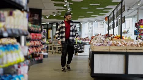 Side-view-of-a-confident-man-in-a-checkered-shirt-and-cap-walking-through-the-supermarket-and-putting-the-products-he-needs-into-a-basket-during-his-shopping