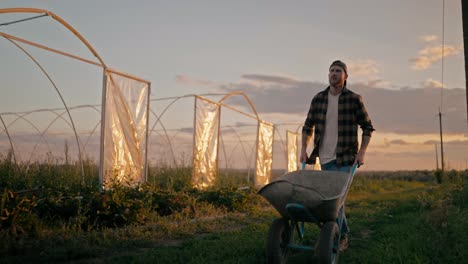 Confident-guy-Farmer-in-a-cap-and-plaid-shirt-rolls-a-wheelbarrow-and-walks-along-a-field-on-a-farm