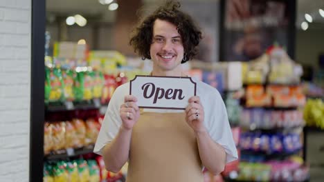 Porträt-Eines-Glücklichen-Brünetten-Kerls-Mit-Lockigem-Haar,-Der-In-Seinen-Händen-Ein-Schild-Mit-Der-Aufschrift-„geöffnet-Nach-Der-Eröffnung-Des-Supermarkts“-Hält