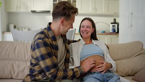 Un-Hombre-Feliz-Con-Una-Camisa-A-Cuadros-Se-Comunica-Con-Su-Esposa-Embarazada-Y-Le-Acaricia-La-Gasa-En-Casa,-En-El-Sofá-De-Un-Apartamento-Moderno.