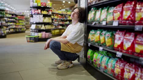 A-tired-guy-with-curly-hair-and-a-yellow-front-supermarket-worker-leaned-on-a-shelf-with-goods-to-rest-during-a-hard-day-at-work