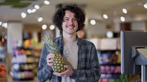 Portrait-of-a-happy-brunette-guy-with-curly-hair-in-a-plaid-shirt-who-holds-a-pineapple-in-his-hands-and-smiles-broadly-in-a-supermarket