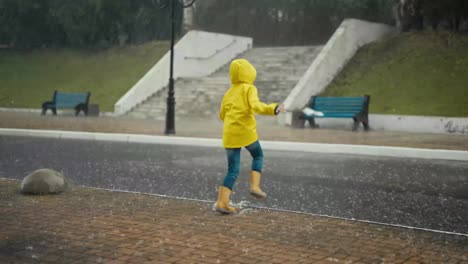 Fröhliches-Teenager-Mädchen-In-Gelber-Jacke-Und-Orangefarbenen-Gummistiefeln-Läuft-Bei-Starkem-Regen-Im-Park-Durch-Große-Pfützen